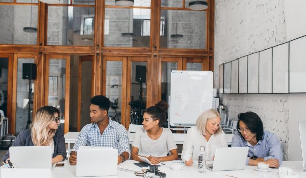 blonde-woman-white-shirt-talking-with-asian-friend-drinking-coffee-near-laptop-with-flipchart-freelance-web-designers-working-together-conference-hall-using-computers-min